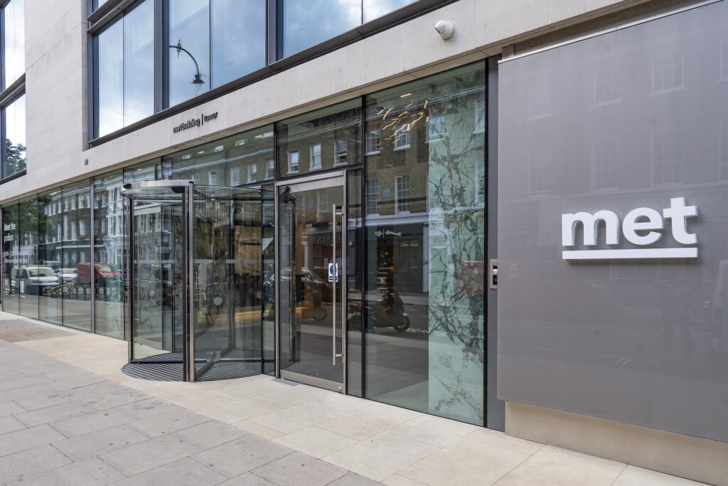 Modern building exterior with large glass windows, a sliding rooflight, and a revolving door entrance. A sign reading "met" is displayed on a grey panel, with street view reflections visible in the glass.