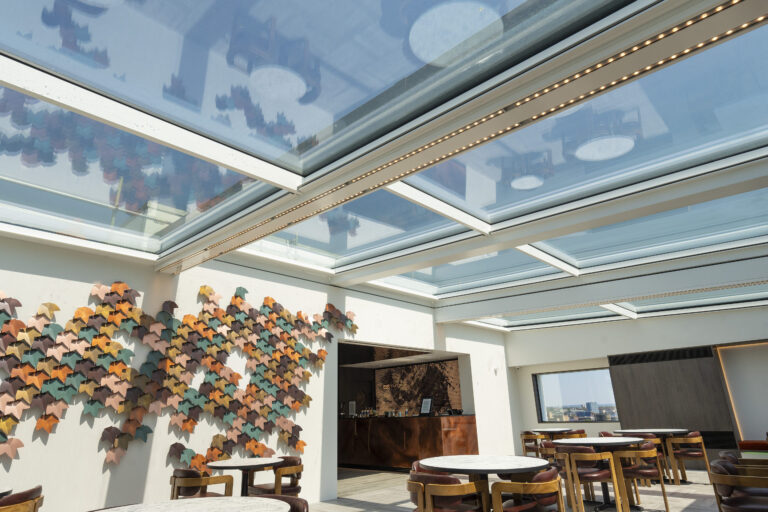 A modern indoor cafe with glass ceilings and artistic wall decor features round wooden tables and chairs arranged for dining. Natural light floods the spacious area.