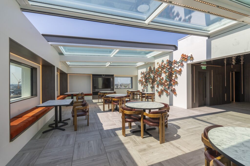 Modern rooftop lounge with large windows, marble-topped tables, wooden chairs, and an art installation on the wall under a glass ceiling featuring a sliding rooflight with stunning city views.