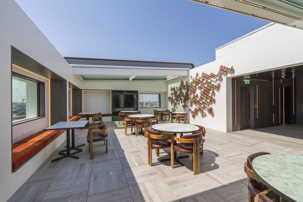 Rooftop lounge with round tables, chairs, window seating, artwork on the wall, and a stunning cityscape view through large windows and a sliding rooflight under a clear sky.