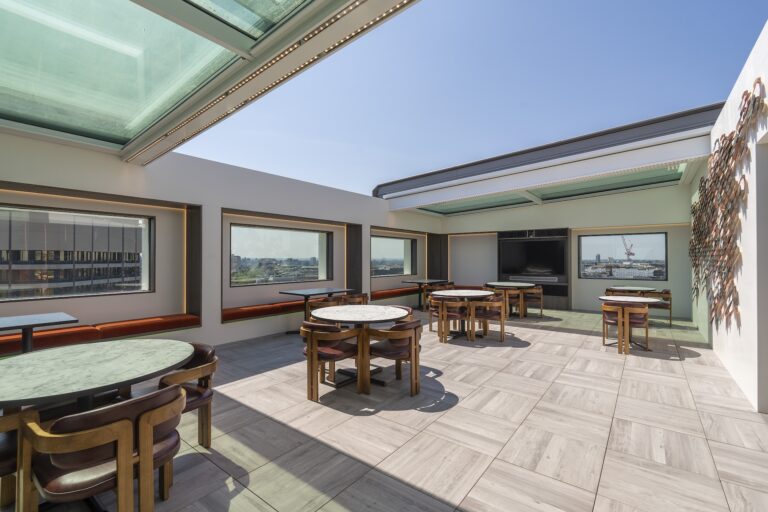 Rooftop dining area with round marble tables, wooden chairs, large windows, and a glass ceiling with a sliding rooflight, overlooking an urban landscape under a clear sky.