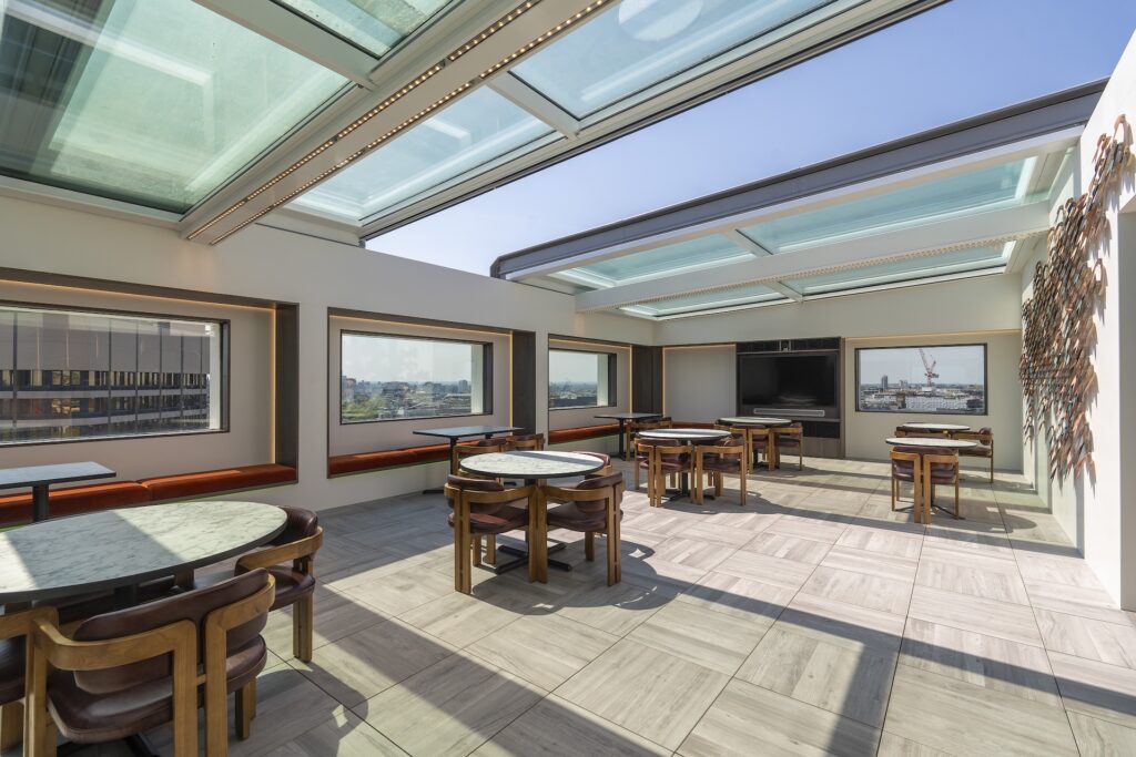 Rooftop dining area with round marble tables, wooden chairs, large windows, and a glass ceiling featuring a sliding rooflight, offering city views in daylight.