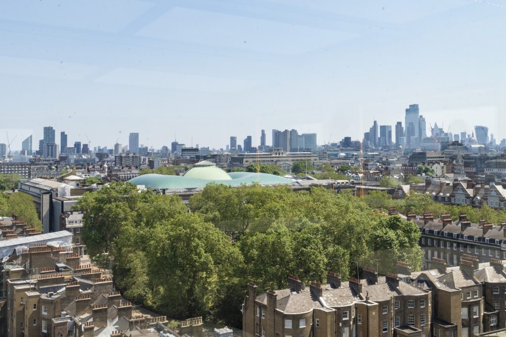 Aerial view of central London with the British Museum’s green roof and sliding rooflight in the foreground, surrounded by trees and buildings, with modern skyscrapers rising in the background.