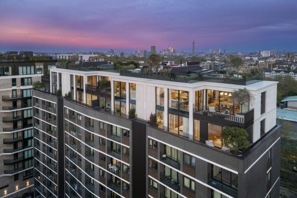 Aerial view of a modern multi-story residential building with balconies and rooftop spaces, set against a cityscape at twilight.