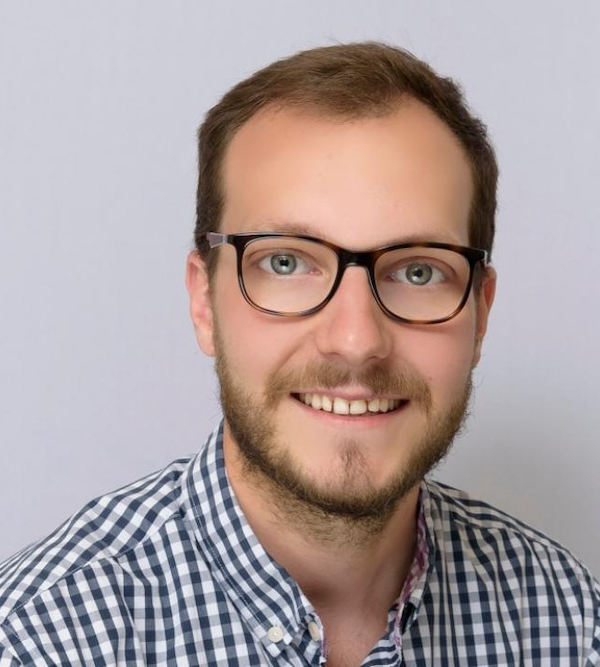 A man with glasses, light brown hair, and a beard is smiling. He is wearing a black and white checkered shirt against a plain background.