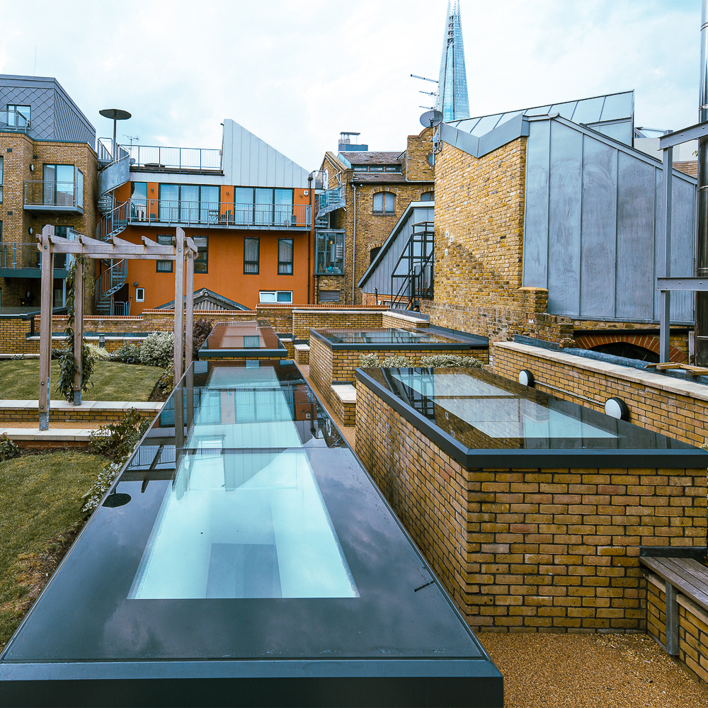 Rooftop view of modern brick buildings with glass skylights and garden areas, set against a cityscape backdrop including the Shard.