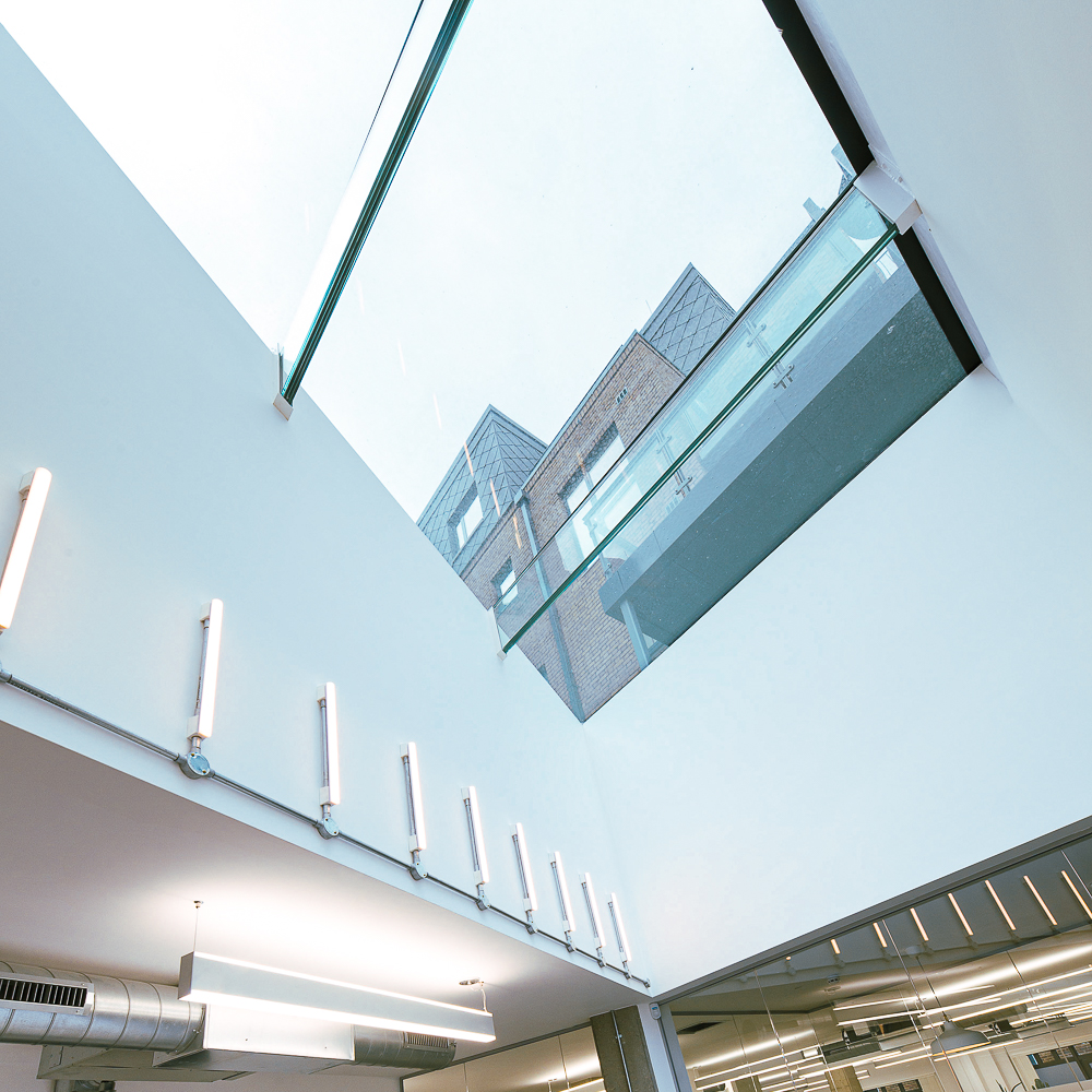 View from inside a modern building with a glass ceiling and walls, revealing an exterior brick structure. Fluorescent lights line the white interior walls.