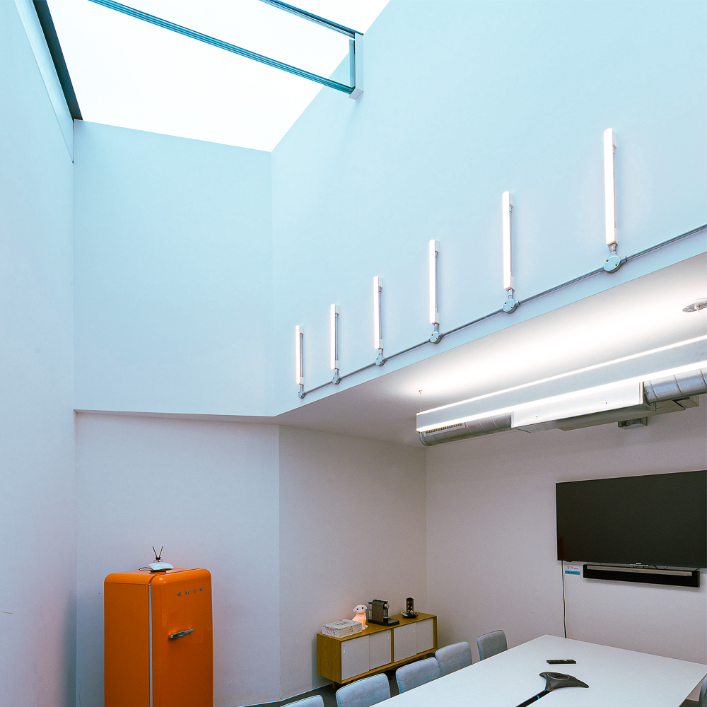 A modern conference room with a long white table, chairs, a wall-mounted TV, an orange fridge, and vertical light fixtures on the walls under a skylight.