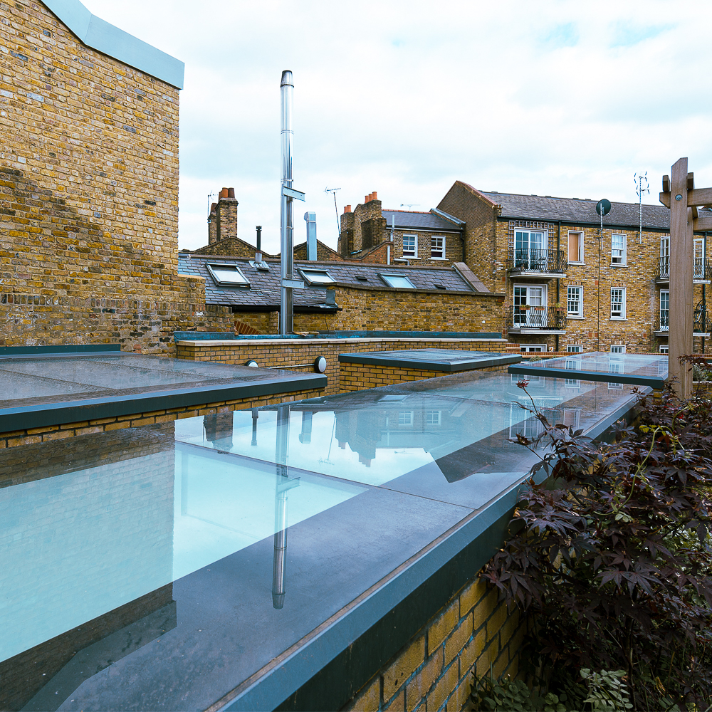 Rooftop with clear glass panels, surrounded by brick buildings and a cloudy sky in the background.