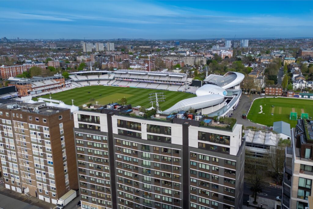 Aerial view of a large cricket stadium with green seating, surrounded by buildings and a cityscape in the background under a blue sky.