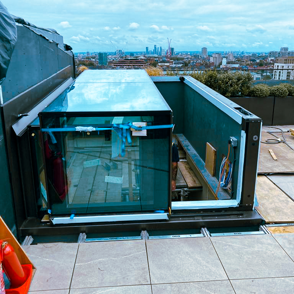 Rooftop view shows a glass skylight structure under construction, surrounded by safety barriers with a city skyline in the background.