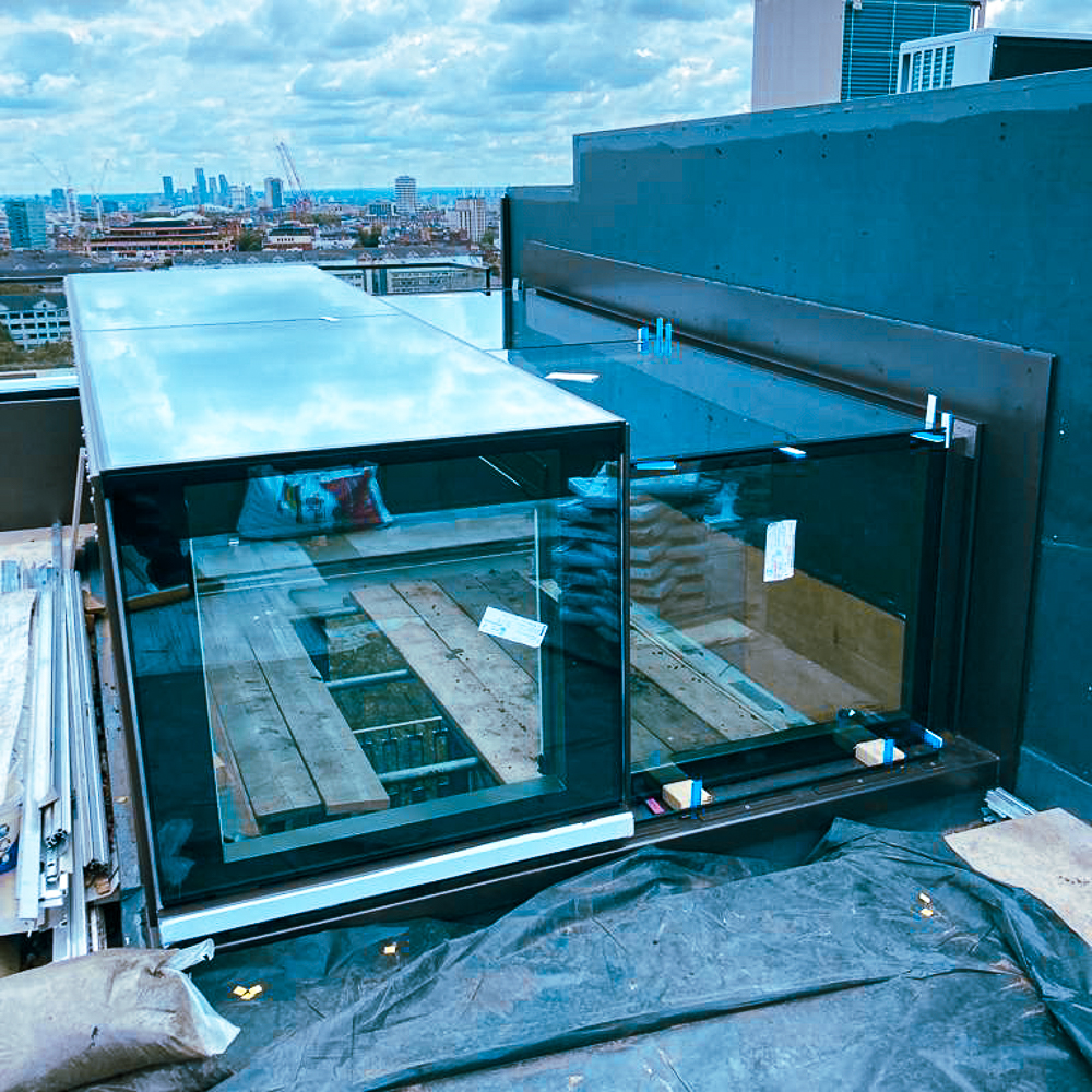A rooftop glass structure expands over the edge, with wooden planks inside. Cityscape and cloudy sky in the background.