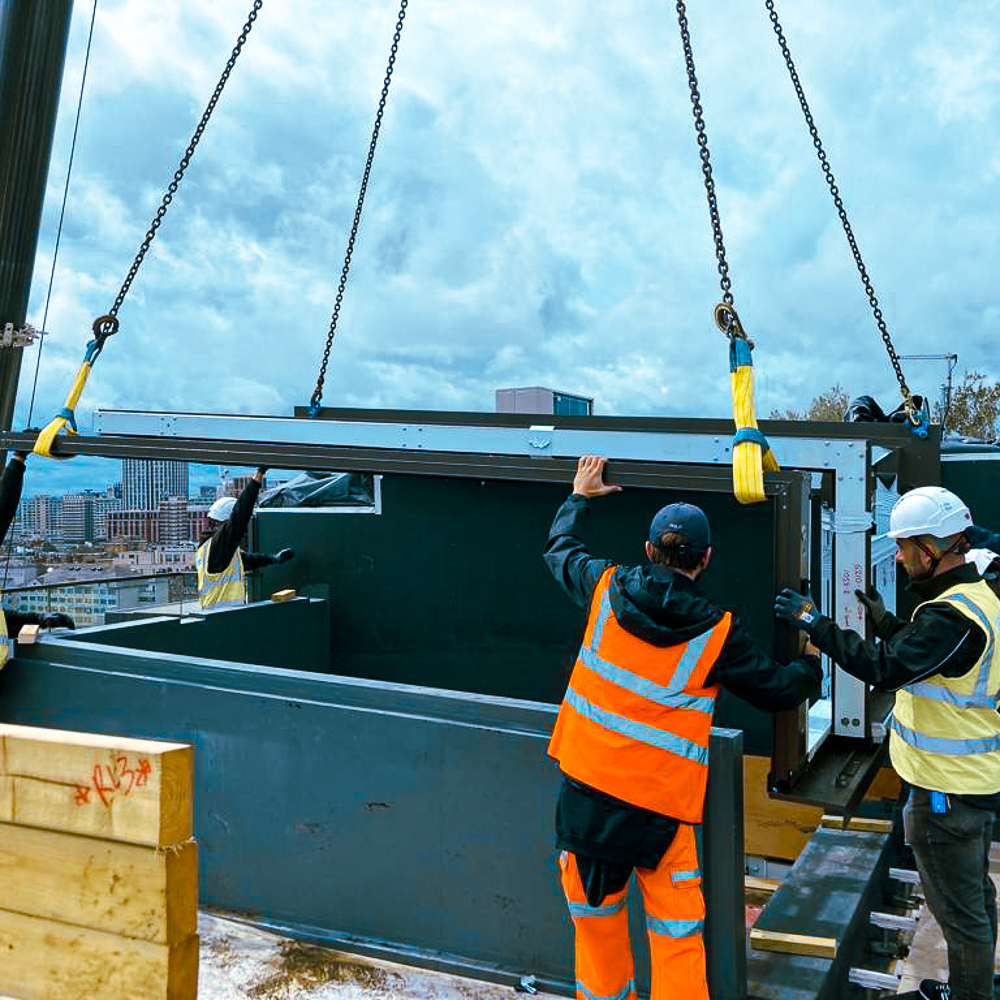 Workers guide a prefabricated unit into place using a crane on a construction site.