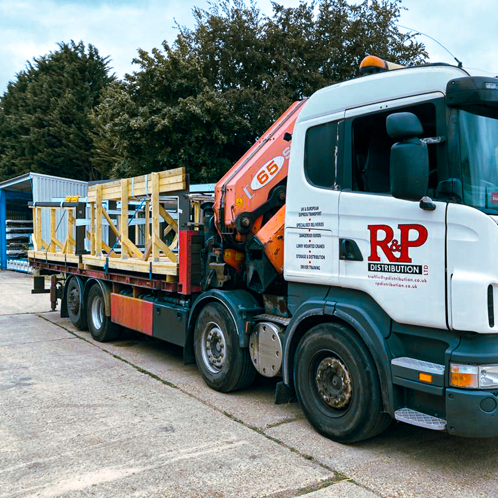 Truck with a side crane, labeled "R & P Distribution," parked, carrying wooden frames. Trees and a metal shed are in the background.