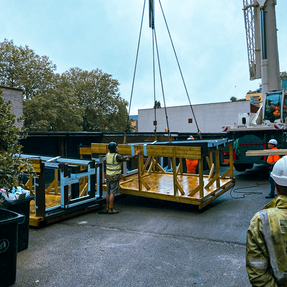 Construction workers are guiding a large metal frame being lifted by a crane on a construction site. Trees and a building are in the background.