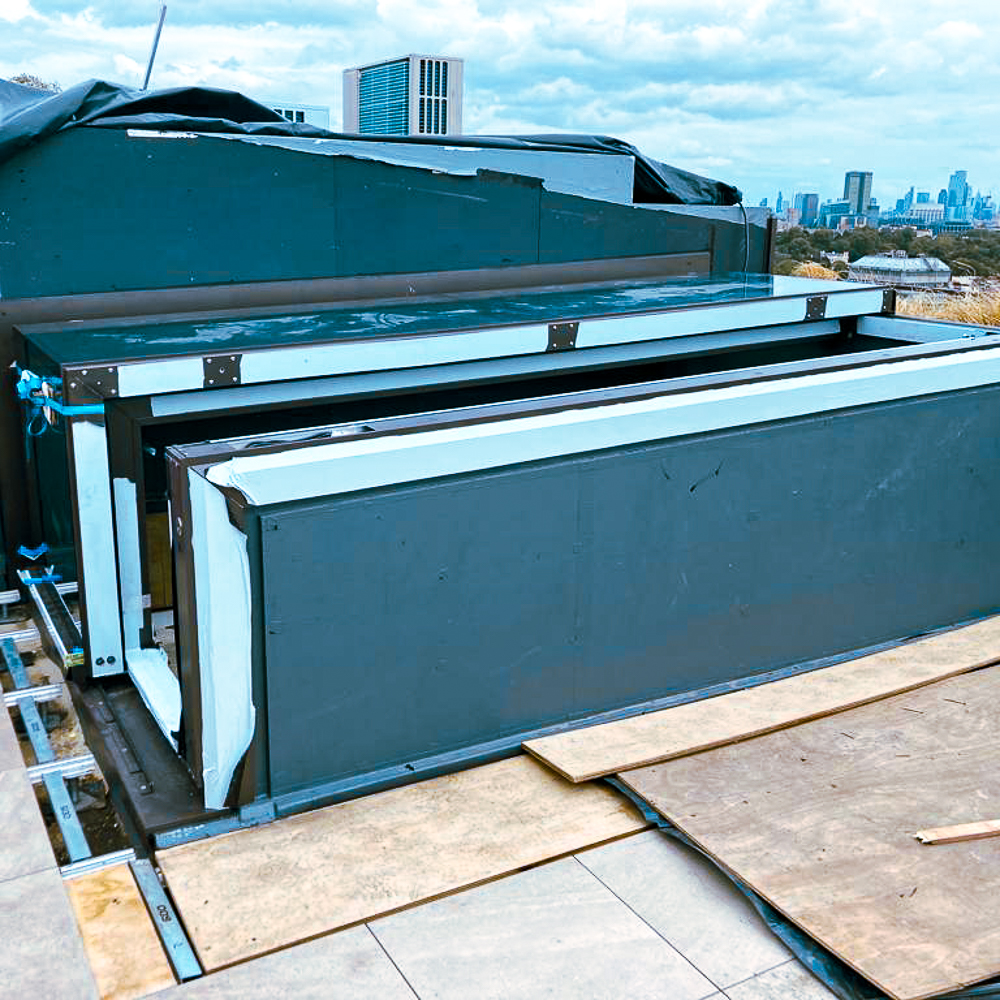 A rooftop with a rectangular metal structure partially covered by a tarp, surrounded by boards, against a cityscape background.