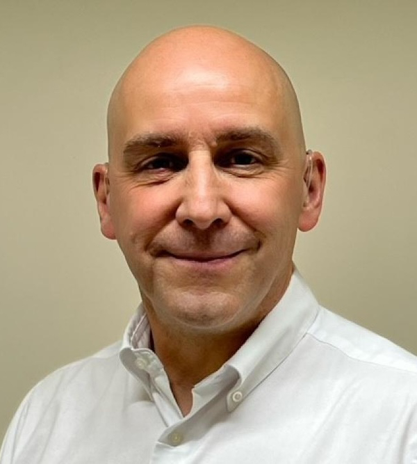 A bald man wearing a white collared shirt smiles in front of a plain background.