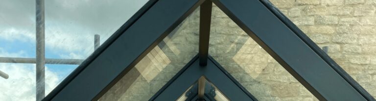 A glass triangle forms a transparent roof structure with metal supports, allowing a view of the cloudy sky and stone wall beyond.