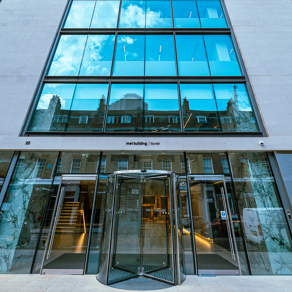 Glass office building facade with revolving door entrance and reflections of clouds and neighboring structures on the windows.