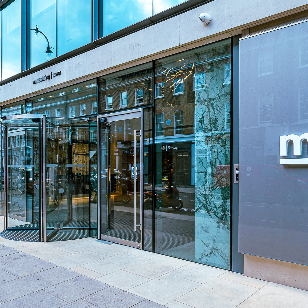 Glass entrance of a modern building with revolving door, street view reflected in glass panels, and nearby motorcycle parked on the street.