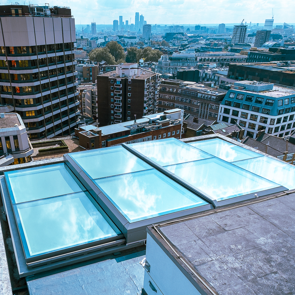 A rooftop with large glass skylights reflects the sky, overlooking a cityscape with various buildings and a blue sky.