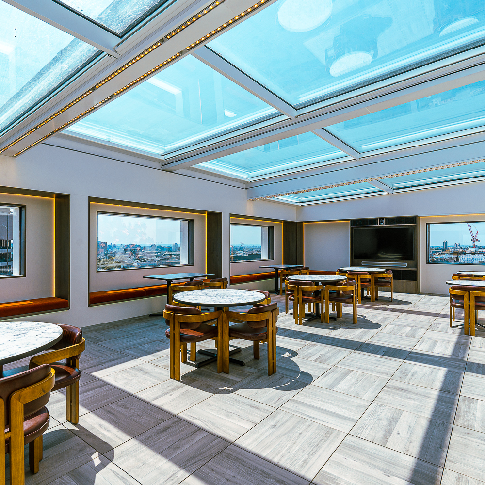 Room with a glass ceiling, round marble tables, and wooden chairs. Large windows provide a panoramic city view.