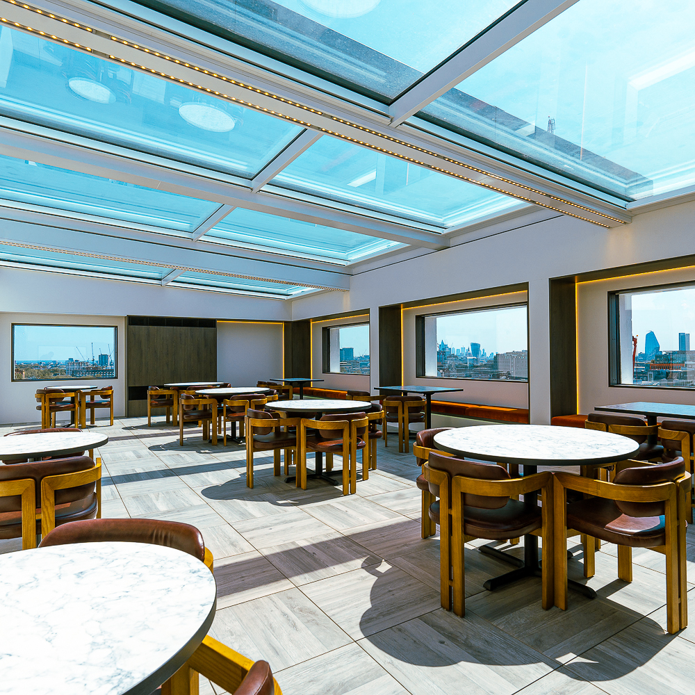 Rooftop dining area with marble tables and wooden chairs, surrounded by large windows offering city views. Skylight ceiling allows natural light.