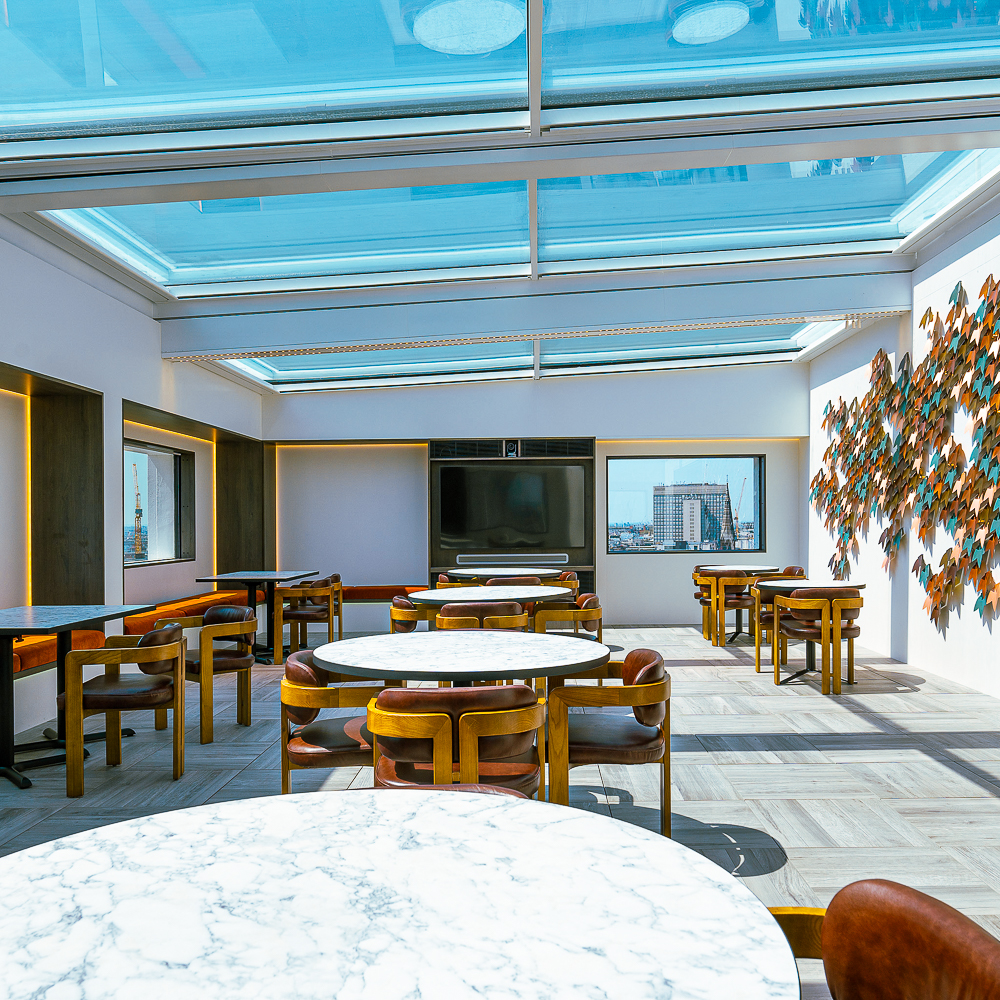 A sunlit room with marble tables, wooden chairs, a wall art piece, and large windows. The glass roof adds brightness to the space.