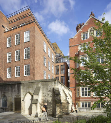 Two multi-story brick buildings with multiple windows stand adjacent to an archway structure. People walk in a courtyard below a partly cloudy sky. Trees are visible on the right.