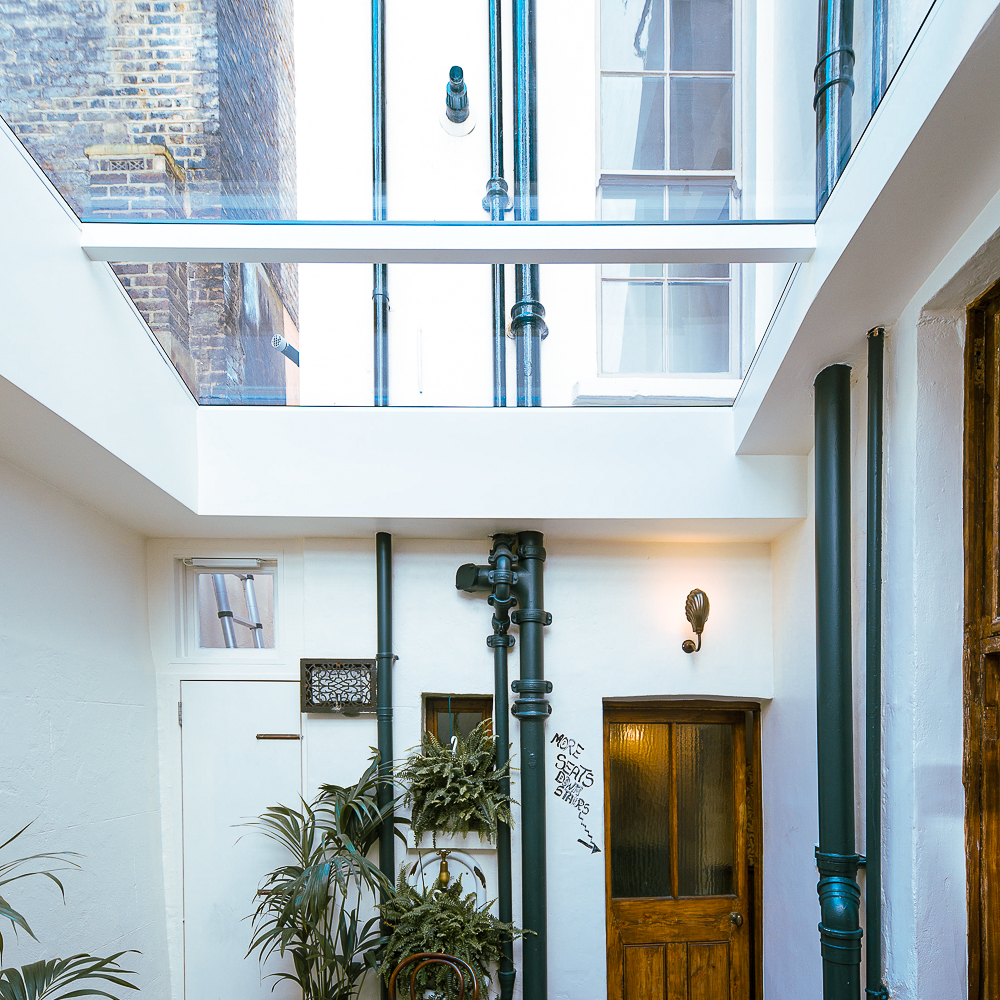 A bright courtyard with white walls, tall green pipes, glass ceiling, and wooden doors. Potted plants are placed against the walls. Graffiti is visible on one wall.