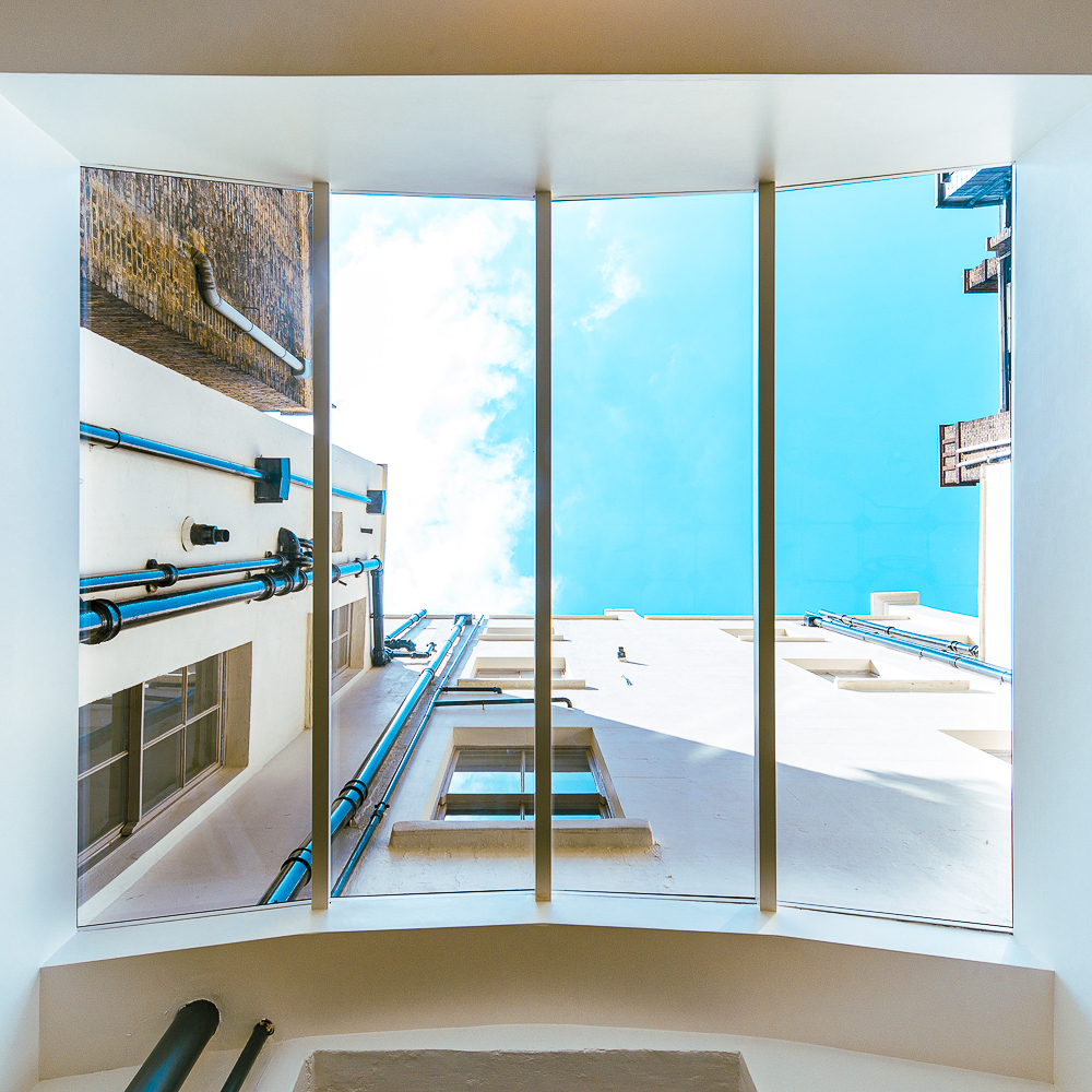 View of a narrow courtyard surrounded by white buildings, looking up toward a blue sky with a few clouds, framed by four vertical window panels.