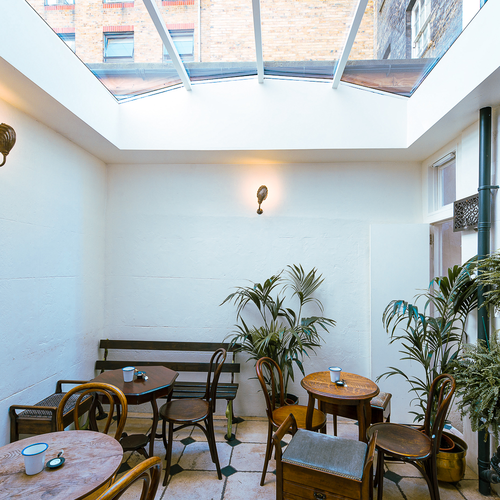 A bright room with a glass ceiling, wooden tables and chairs, potted plants, and a wall light.
