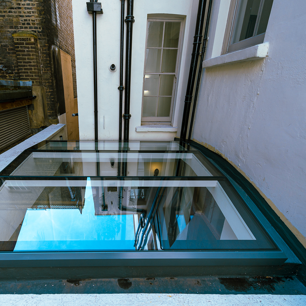 A glass skylight on a flat rooftop reflects the surrounding buildings and sky, with adjacent walls, windows, and pipes in view.