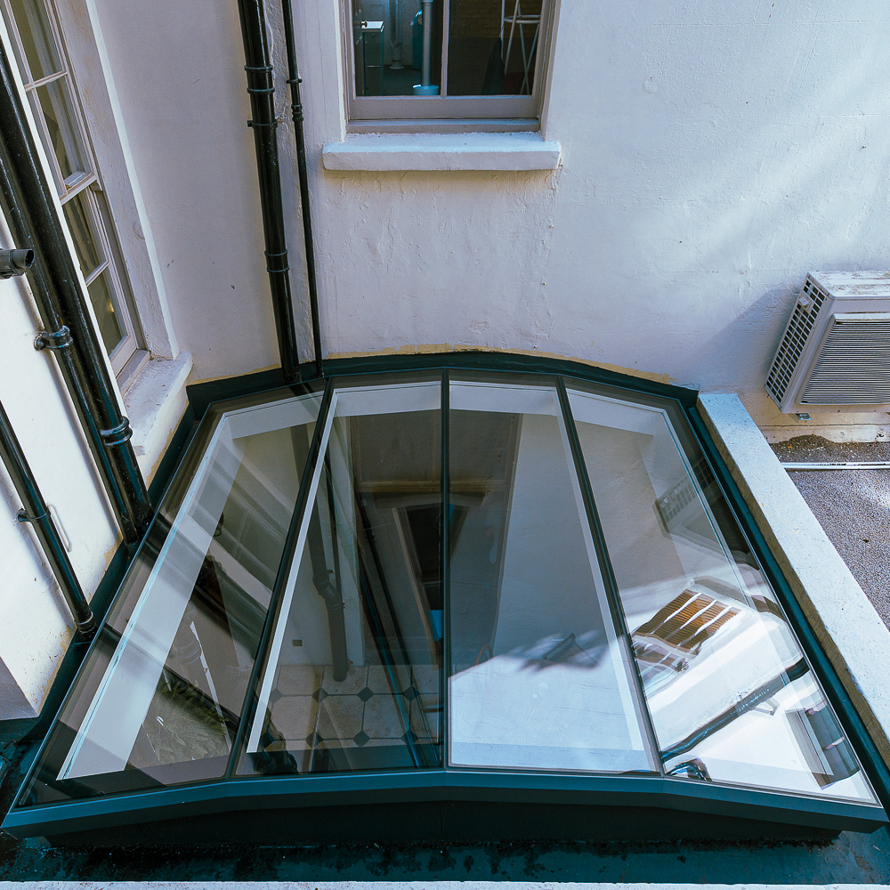 Glass skylight with multiple panels installed on a flat roof, surrounded by white walls and adjacent to windows and a wall-mounted air conditioning unit.