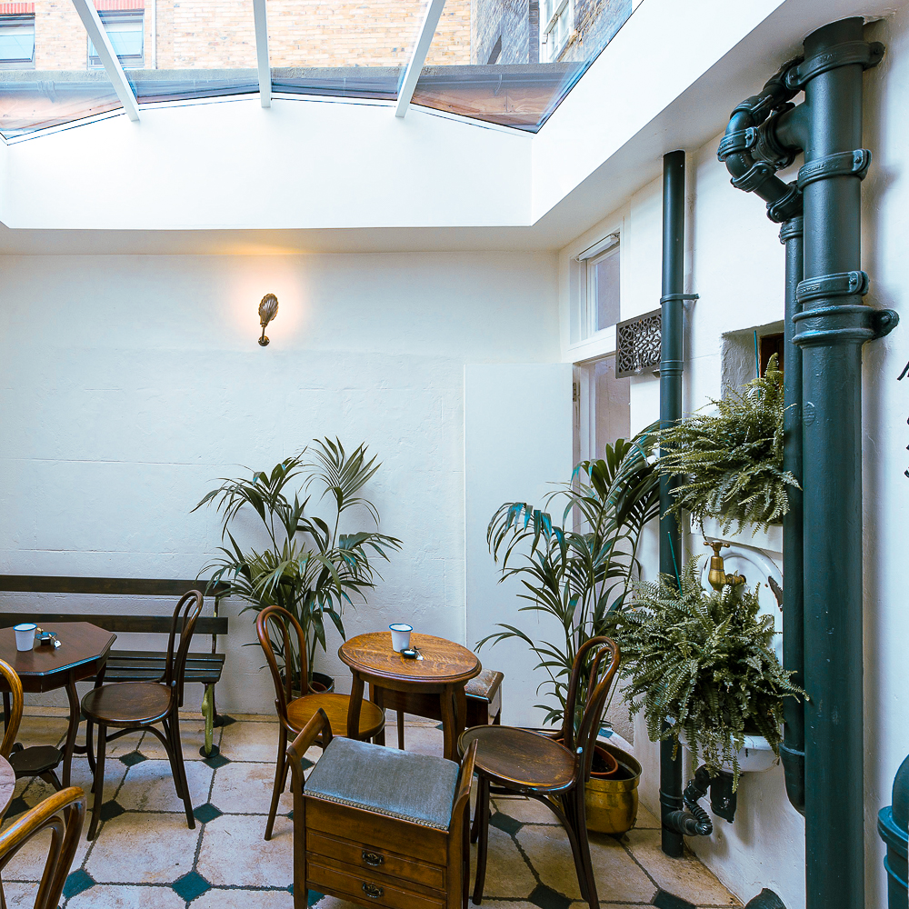Indoor seating area with wooden chairs and tables, potted plants, and a skylight ceiling. Walls are white, with a mix of natural light and a wall lamp.