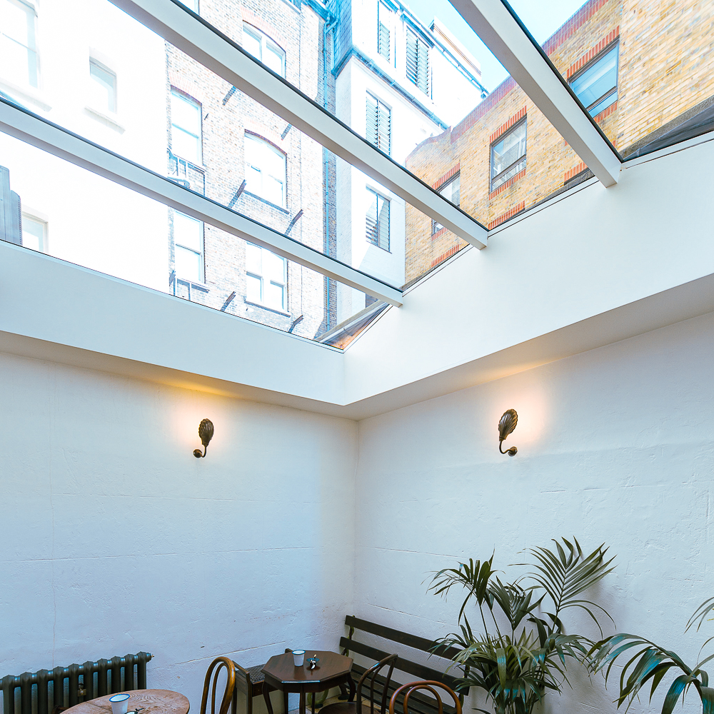 Interior of a room with a glass ceiling, white walls, and two wall lamps. There are wooden chairs, a small table, and a plant in the corner. Several buildings are visible through the ceiling.