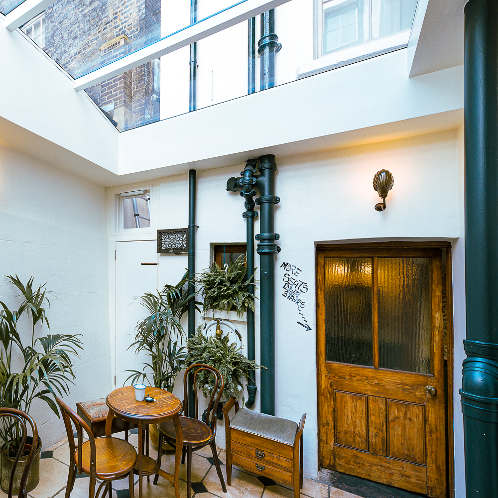 A small courtyard with plants, a wooden table and chairs, a glass ceiling, and a door next to graffiti on the wall.
