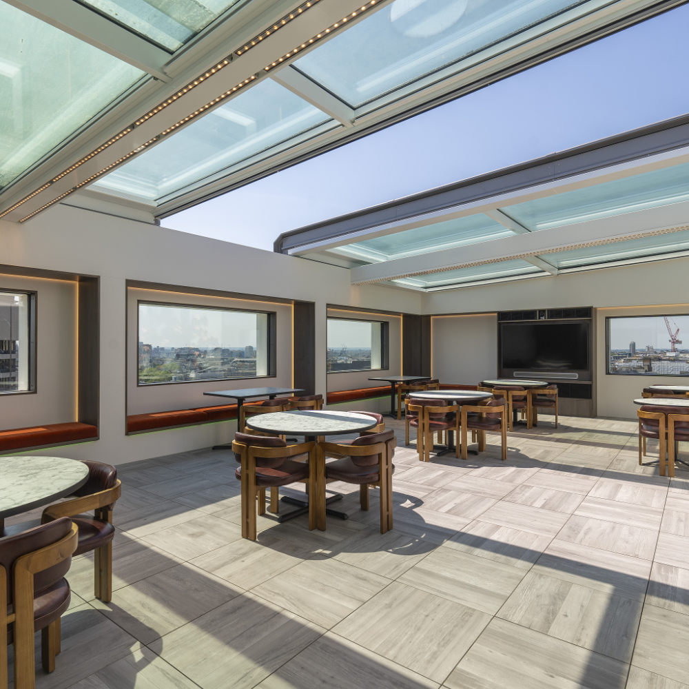 NGR Bi Part Electric Opening Sliding Rooflight Partially Open A spacious, sunlit room features several round tables and chairs under a large, retractable glass roof with tile flooring.