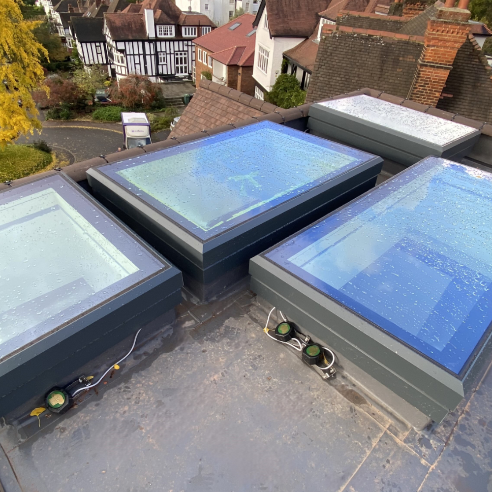 NGR Electric Opening Rooflights With Sensors Rooftop view showing several rectangular skylights with rain droplets on them, surrounded by a residential neighborhood with houses and trees.