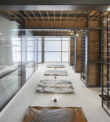 Room with large windows and a high ceiling featuring several metal-framed beds with brown covers. The space has an industrial design with exposed beams and a minimalist aesthetic.