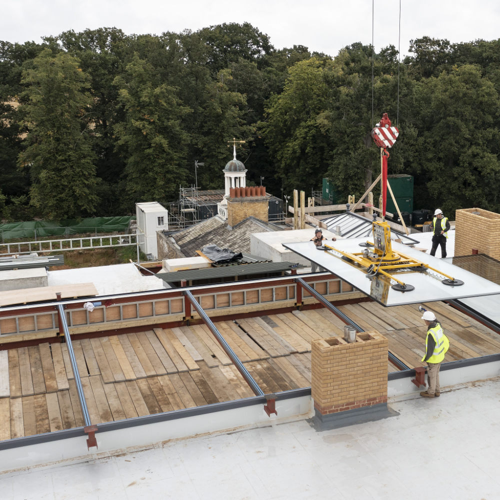 NGR High End Residential Two Ton Elevated Rooflight installation via Crane Construction workers install large glass panels using a crane on a rooftop, surrounded by greenery and scaffolding on nearby structures.