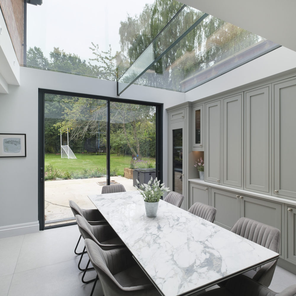 A modern kitchen and dining area with a large glass skylight, marble dining table, and sliding glass doors leading to a garden with a soccer goal and trees in the background.