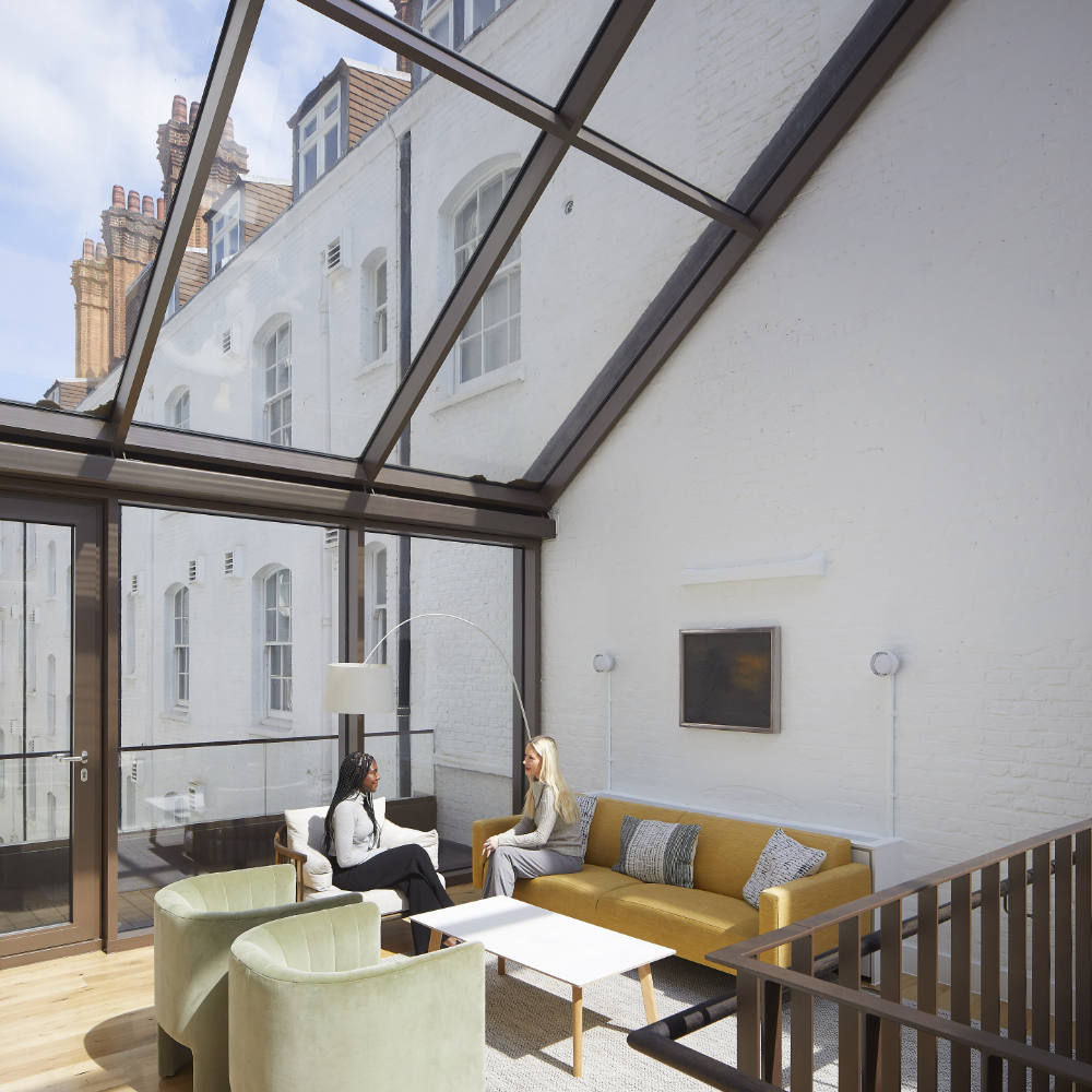 NGR Pitch Multi Panel Fixed Rooflights Commercial Project London Two women sit and converse on a yellow couch under a glass ceiling in a modern, well-lit room with white walls and contemporary furniture.