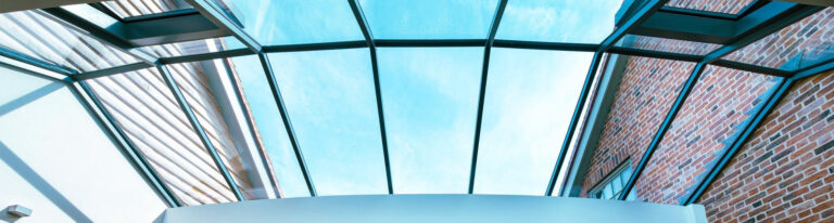 Glass ceiling structure with black metal frames, providing a view of a clear blue sky. The surrounding building walls are made of brick and metal.
