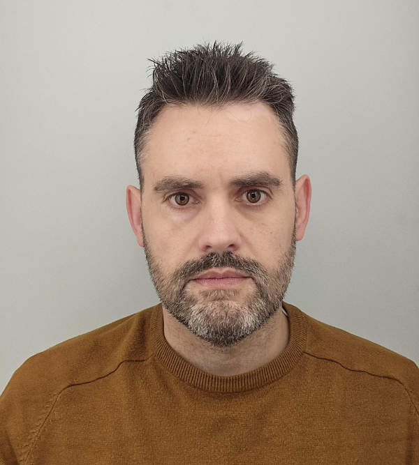 A man with short, dark hair and a trimmed beard wearing a brown sweater stands against a plain, light-colored background, looking directly into the camera.