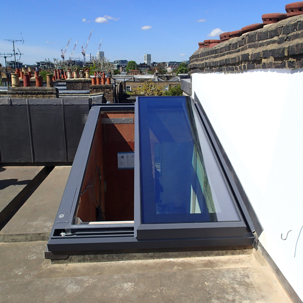 Snow-covered flat rooftop with a square partially finished skylight opening. Nearby, there's a blue sky and distant trees visible.