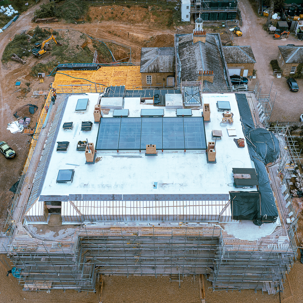 Aerial view of a construction site with scaffolding surrounding a large building. The roof is partially covered, and construction materials are visible.