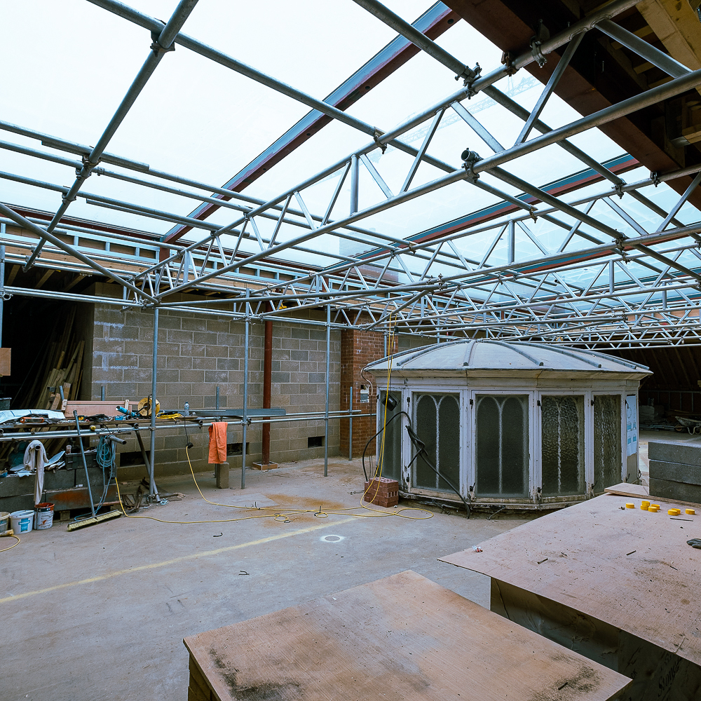 Construction site with metal scaffolding, tools, and an octagonal skylight structure on the floor. Walls are unfinished, and the ceiling framework is visible against the light.