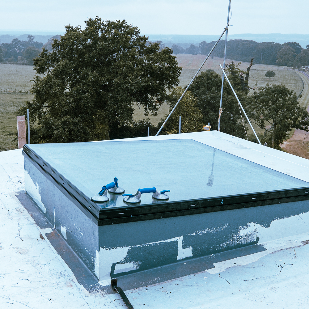A glass rooftop hatch with blue sealant and suction cups, set against a landscape of trees and fields in the background.