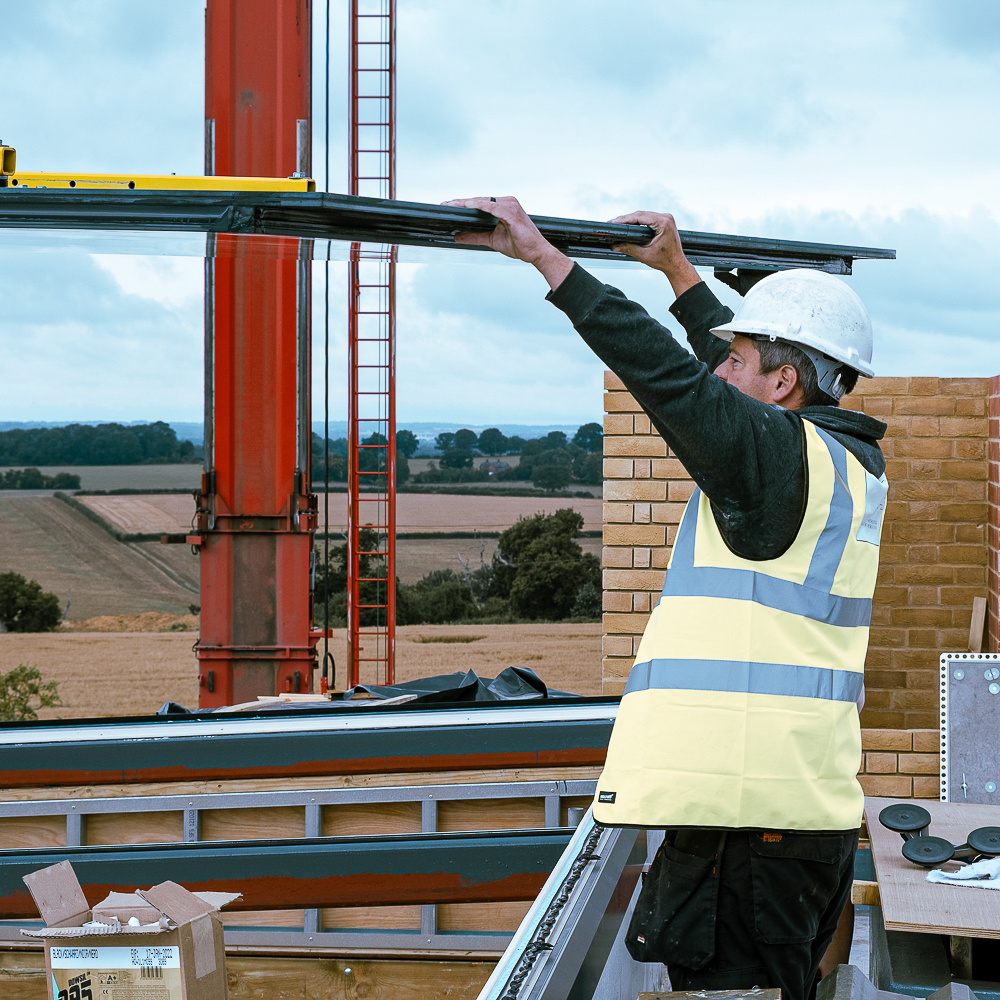 Construction worker in a hard hat and high-visibility vest installing a panel on a building with a crane in the background.
